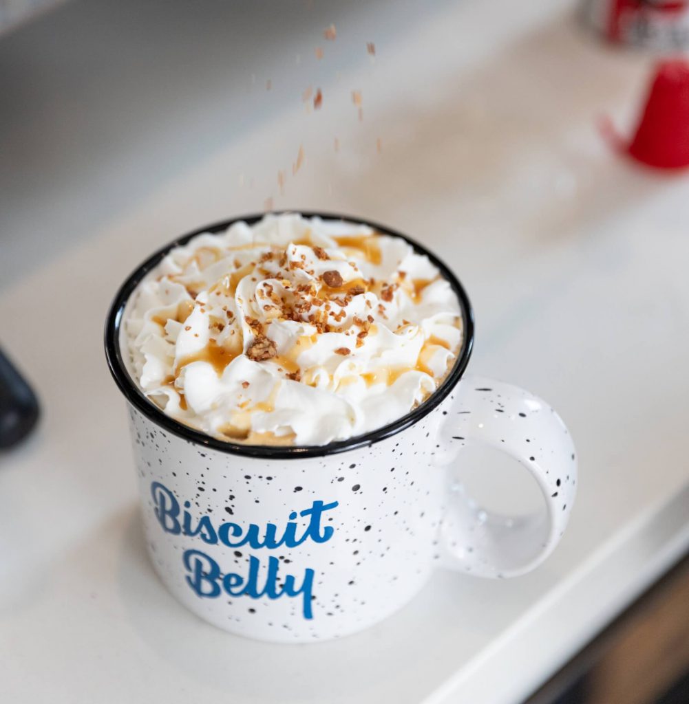 Picture of praline bits being sprinkled on a praline mocha with whip cream and caramel in a Biscuit Belly branded mug