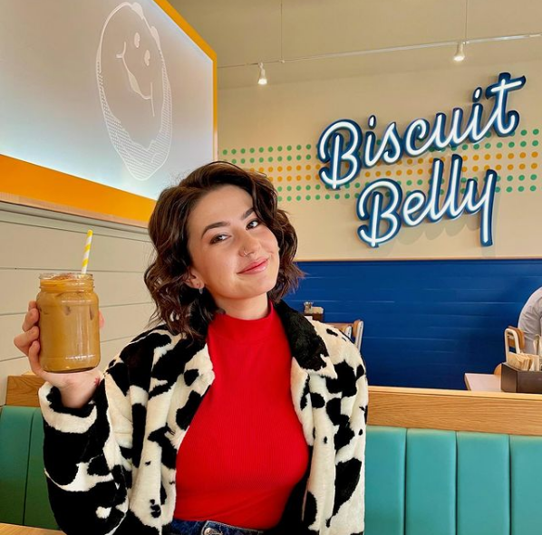 Brunette woman in a red top and printed jacket holds a glass of iced coffee in front of a neon Biscuit Belly sign