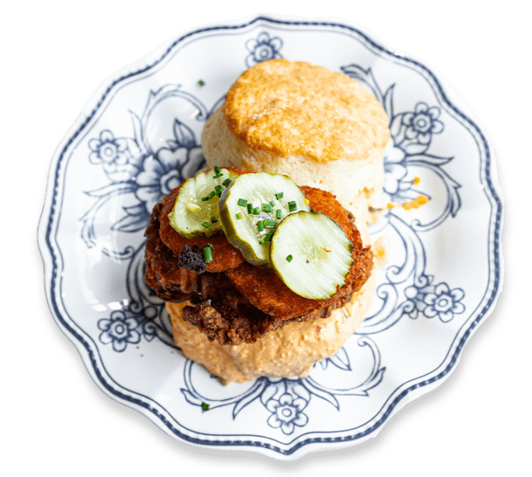 Picture of an open-faced biscuit sandwich with pickles, fried green tomatoes, and fried chicken on a blue and white plate
