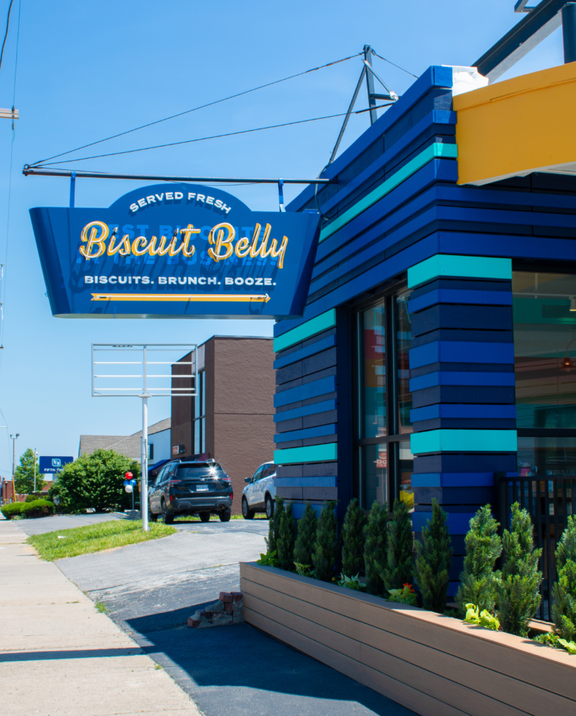 The storefront of Biscuit Belly in the Grandview Heights neighborhood of Columbus Ohio, a navy and teal striped facade with a vintage inspired neon Biscuit Belly sign that says Biscuits, Brunch & Booze.