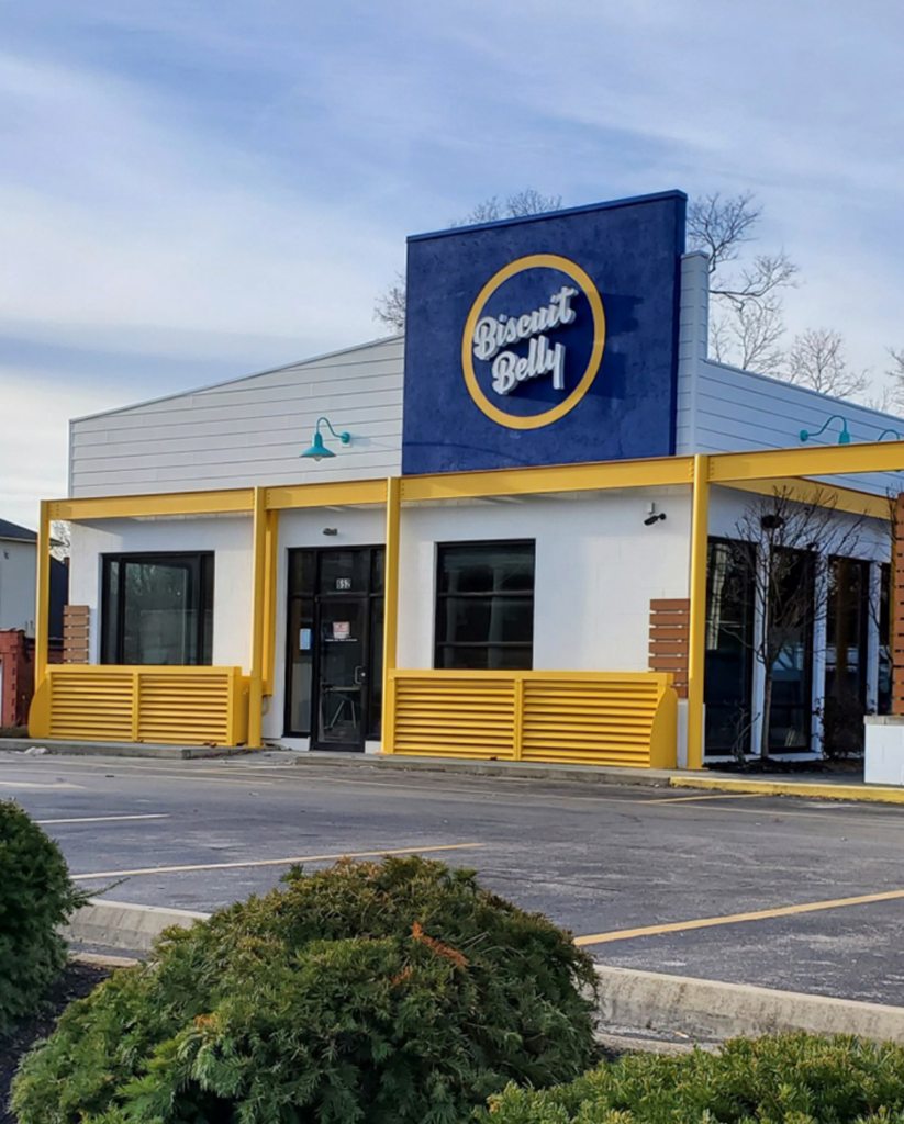The front side of the Biscuit Belly Lexington store location. A white building with yellow accents, teal gooseneck lamps and the Biscuit Belly logo in Navy, White & yellow.