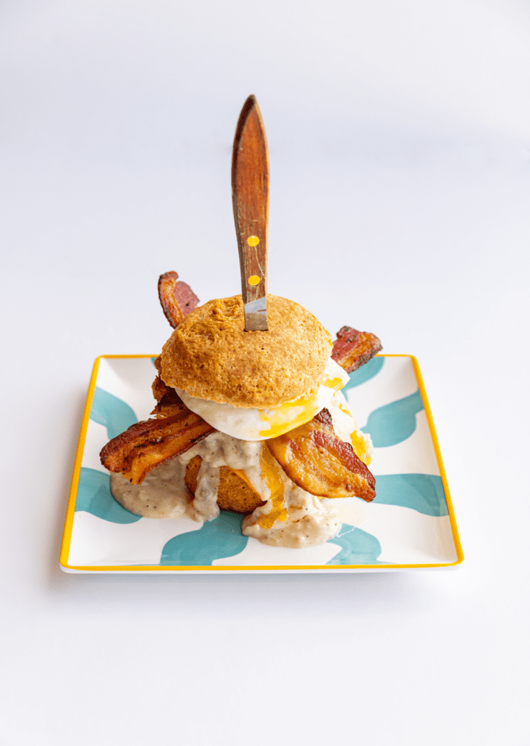 Picture of a biscuit sandwich with cheese, bacon with a knife stuck through it on a blue and white plate