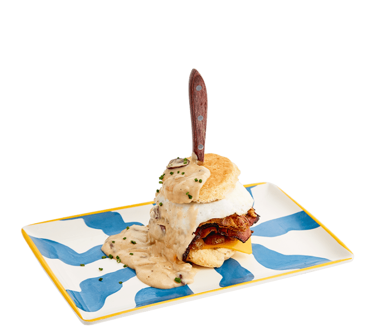 a biscuit sandwich stacked with every type of protein: smoked brisket, sausage, bacon, fried chicken, cheddar cheese, an over easy egg and topped with mushroom gravy, stabbed through the top with a wooden handled knife on a blue and white striped plate.