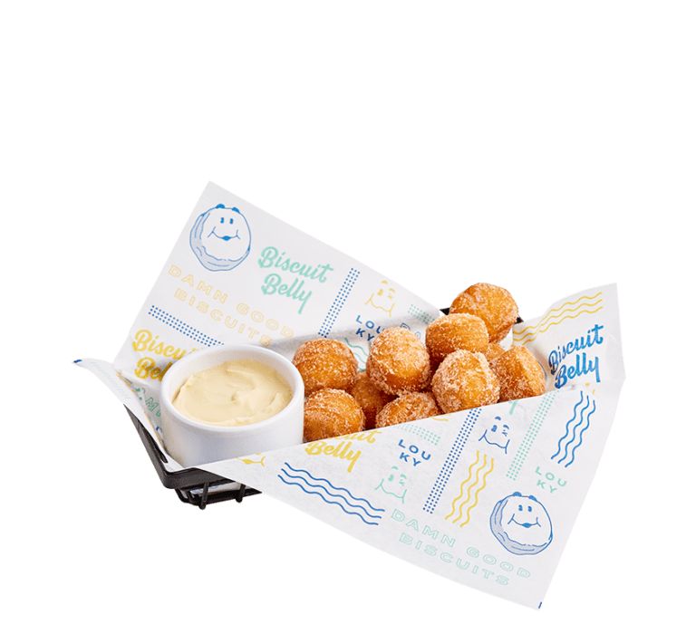 black wire basket lined with biscuit belly branded paper and filled with sugar and cinnamon coated fried donut holes with a side of frosting in a small white bowl