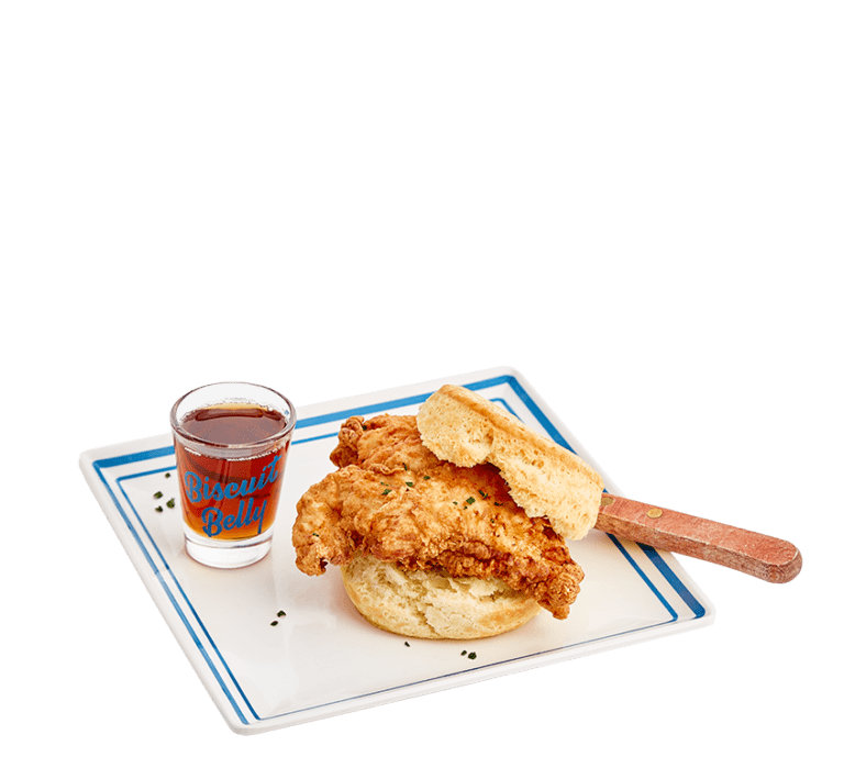 a biscuit sandwich topped with fried chicken with a wooden handled knife under the biscuit top and a biscuit belly branded shot glass filled with bourbon maple syrup on a white plate with a blue border