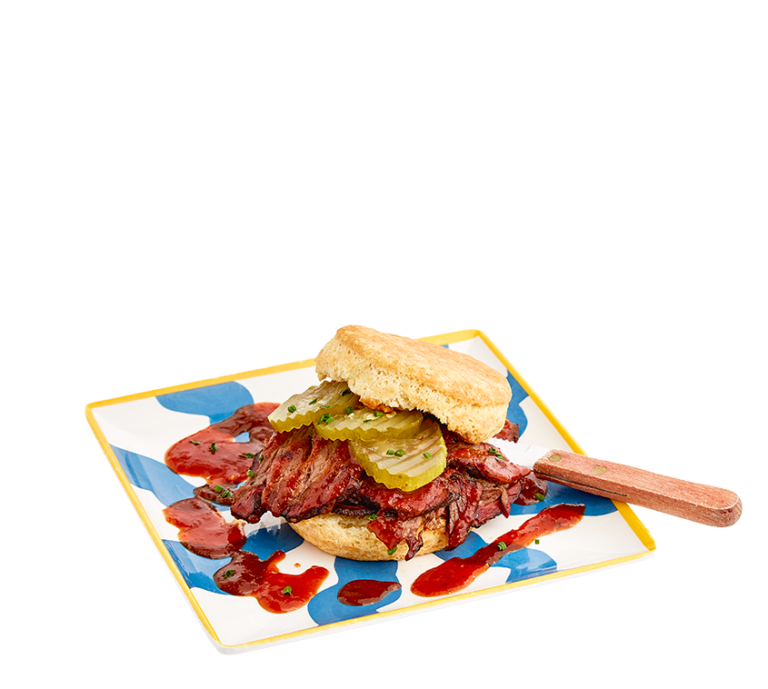 a biscuit sandwich made with smoked brisket, topped with a slightly spice, red poblano-tomato sauce and three pickles, served on a colorful blue and white striped plate and a wood-handled knife tucked under the biscuit top.