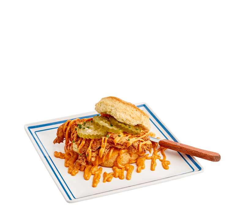 a biscuit sandwich with crispy fried chicken, drizzled with a hot nashville mayo sauce, three pickles on a white plate with blue border and a wooden handled knife tucked under the biscuit lid