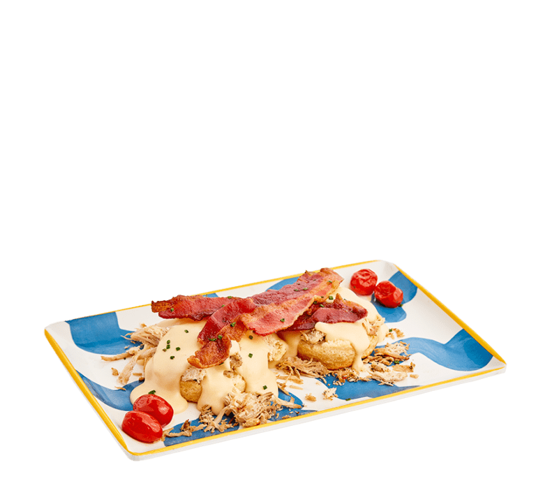 an open faced biscuit topped with shredded turkey, smoked gouda mornay gravy, two slices of thick-cut bacon and two roasted cherry tomatoes on two corners of a blue and striped rectangular plate.
