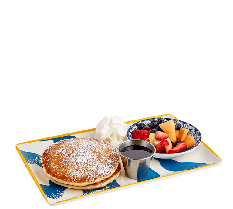 two pancakes topped with powdered sugar, mixed fruit in a small blue and white bowl, whipped cream, and syrup in a silver metal ramekin on a blue and white plate with a yellow border