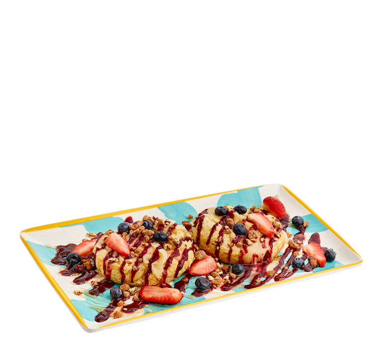 an open faced biscuit topped with melted brie, berry jam, honey, strawberries, blueberries, and crushed pralines on a teal and white rectangular plate with a yellow border