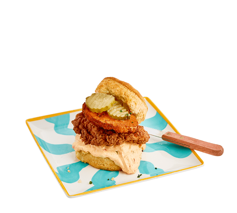 a biscuit sandwich made with crispy, buttermilk fried chicken, a light orange pimento cheese that's peppered with jalapeno, topped with a fried green tomato and pickles on a teal and white striped square plate.
