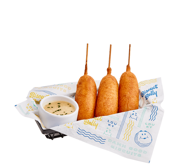 a black wire basket lined with a Biscuit Belly branded paper filled with three corndogs served with a side of sweet maple mayo in a small white bowl.