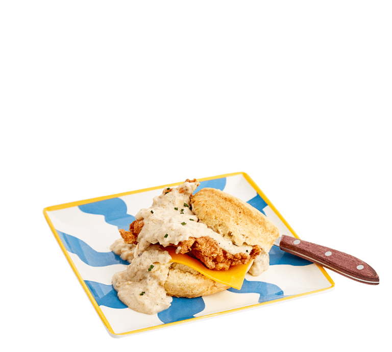 a biscuit sandwich topped with cheese, fried chicken, and sausage gracy on a blue and white plate with a yellow border and a wooden handled knife tucked under the biscuit top