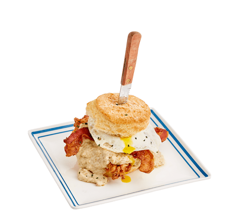 a biscuit sandwich topped with cheese, fried chicken, sausage gravy, bacon, and an over easy egg with a wooden handled knife stabbed through it on a white plate with a blue border