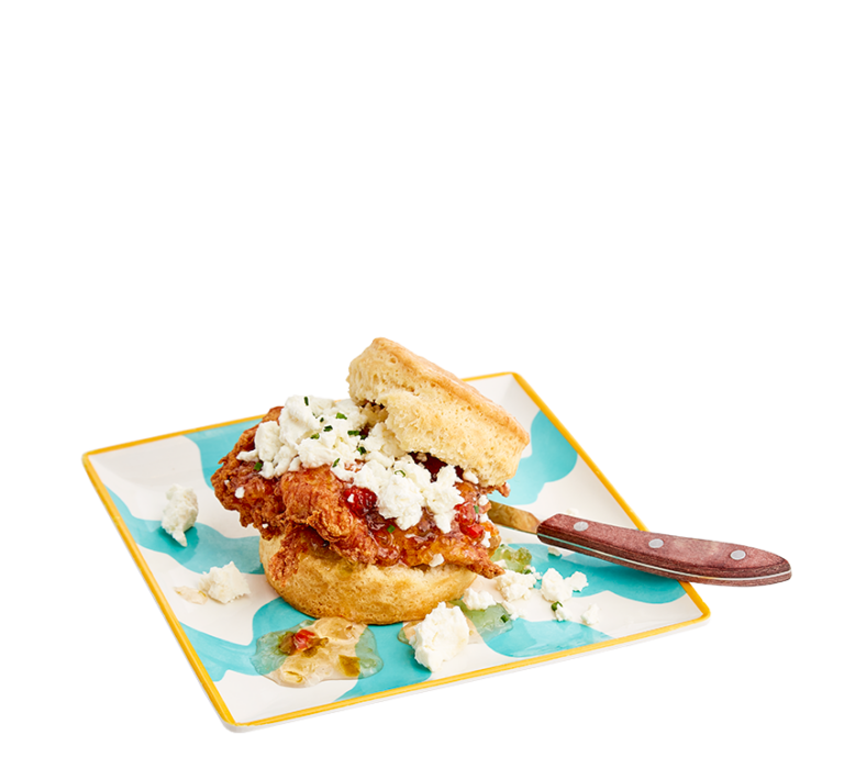 A biscuit sandwich topped with fried chicken, pepper jelly, and goat cheese on a teal and white square plate with a wooden handled knife under the biscuit top.