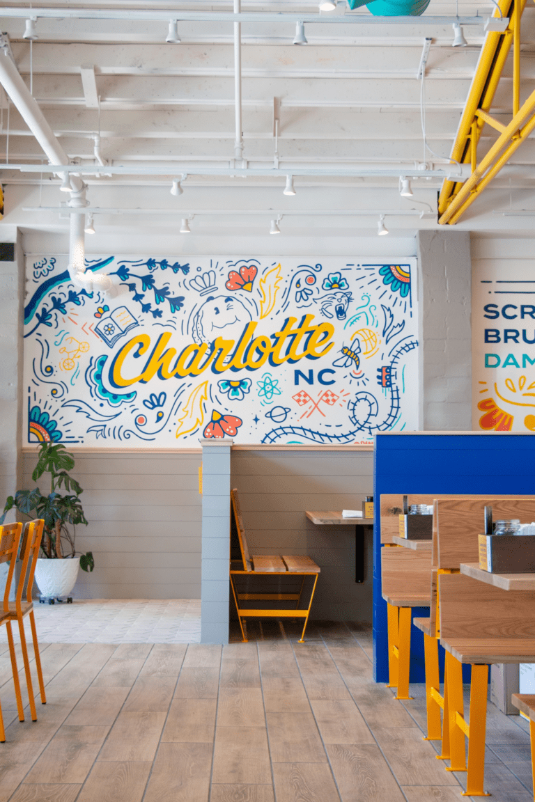 An interior image of Biscuit Belly location in Charlotte, NC with a colorful mural that reads "Charlotte, NC" in script letters and surrounded by colorfully painted, local callouts.
