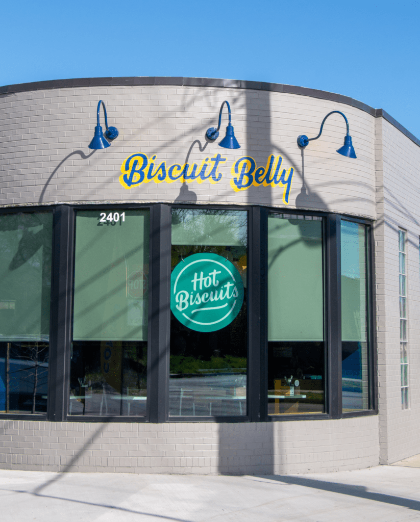 An image of the exterior face of Biscuit Belly in Norfolk with a gray, painted brick, Biscuit Belly logo painted in a blue and yellow script with three gooseneck lights and a teal Hot Biscuits sign hanging in the window.