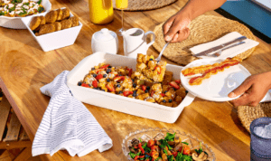 A young man uses a silver spatula to serve himself a piece of french toast bake from a white casserole dish onto a white plate with two pieces of bacon.