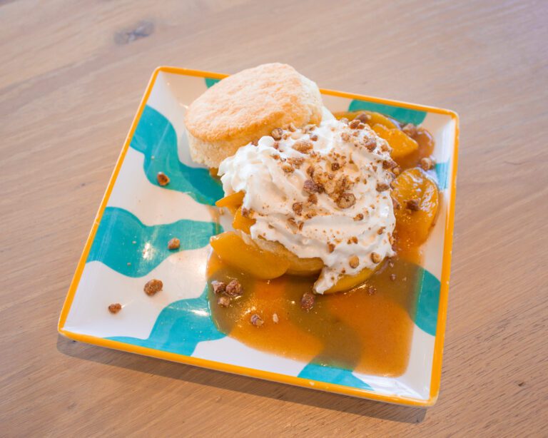 A scratch-made buttermilk biscuit topped with a bourbon caramelized peach topping and whipped cream and crushed pralines on a blue and white plate sitting om a light wooden table.