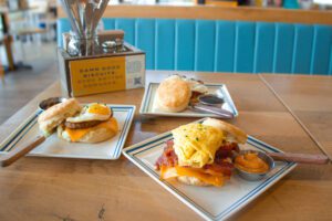 Three homemade buttermilk biscuits on white plates, outlined in blue sit on a light brown wooden table. The closest biscuit sandwich features a scrambled egg, bacon, and cheddar cheese. A small silver cup is places on the right side of the sandwich with our homemade sauce.