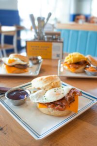 Three homemade buttermilk biscuit sandwiches on white square plates, outlined in blue, sit on a light brown wooden table. The closest biscuit sandwich features an egg, bacon, and cheddar cheese. A small silver cup of dark purple berry jam is to the left. The two blurred biscuit sandwiches in the background have a silverware and napkin holder behind them.