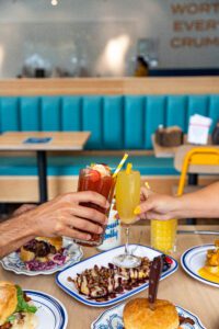 Two hands clink cups together in a cheerful toast over a table filled with brunch treats. The table features biscuit sandwiches, open faced love shack covered in honey and berry jam, and the signature Biscuit Belly Rockwell, with a knife pierced into the sandwich to secure it together. 