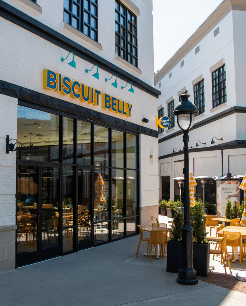 The front entrance of Biscuit Belly Peachtree city, located in the avenue. A white painted brick storefront with teal gooseneck lights and a BISCUIT BELLY sign in Blue & Yellow lettering.
