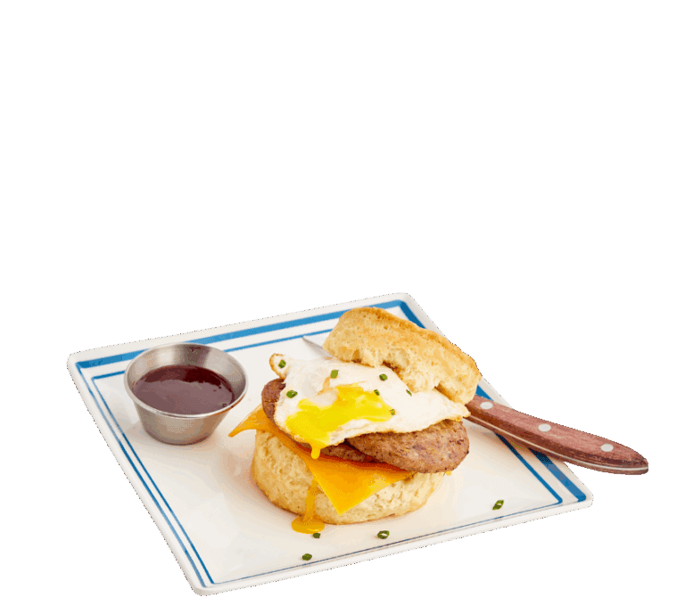A biscuit sandwich with sausage patties, cheddar cheese, and a runny egg on a square plate with a blue border. A small, silver ramekin sits on the plate next to the sandwich with a wooden handled knife tucked under the biscuit lid.