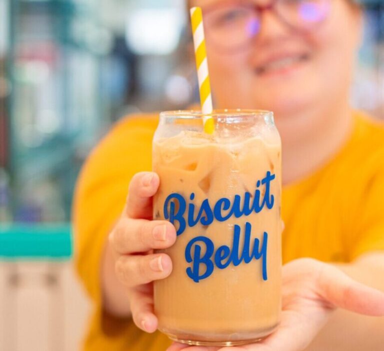 A server holds an iced latte in a glas with the Biscuit Belly logo in navy printed on it with a yellow and white striped straw.