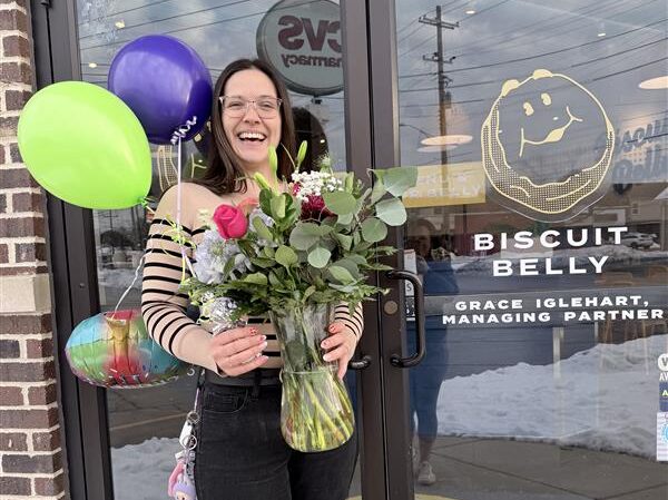Grace stands in front of Biscuit Belly St Matthew's entrance with her name on the door, holding balloons and flowers to celebrate this milestone.
