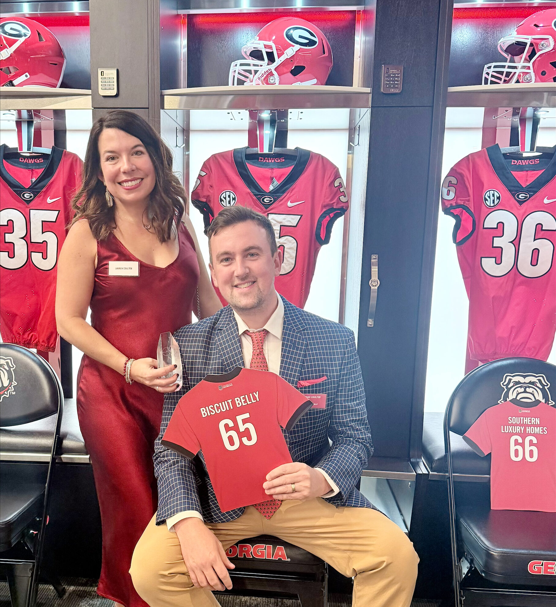 Chad & Lauren Coulter in the UGA Football locker room holding a jersey shaped plaque that says "BISCUIT BELLY 65" on it.